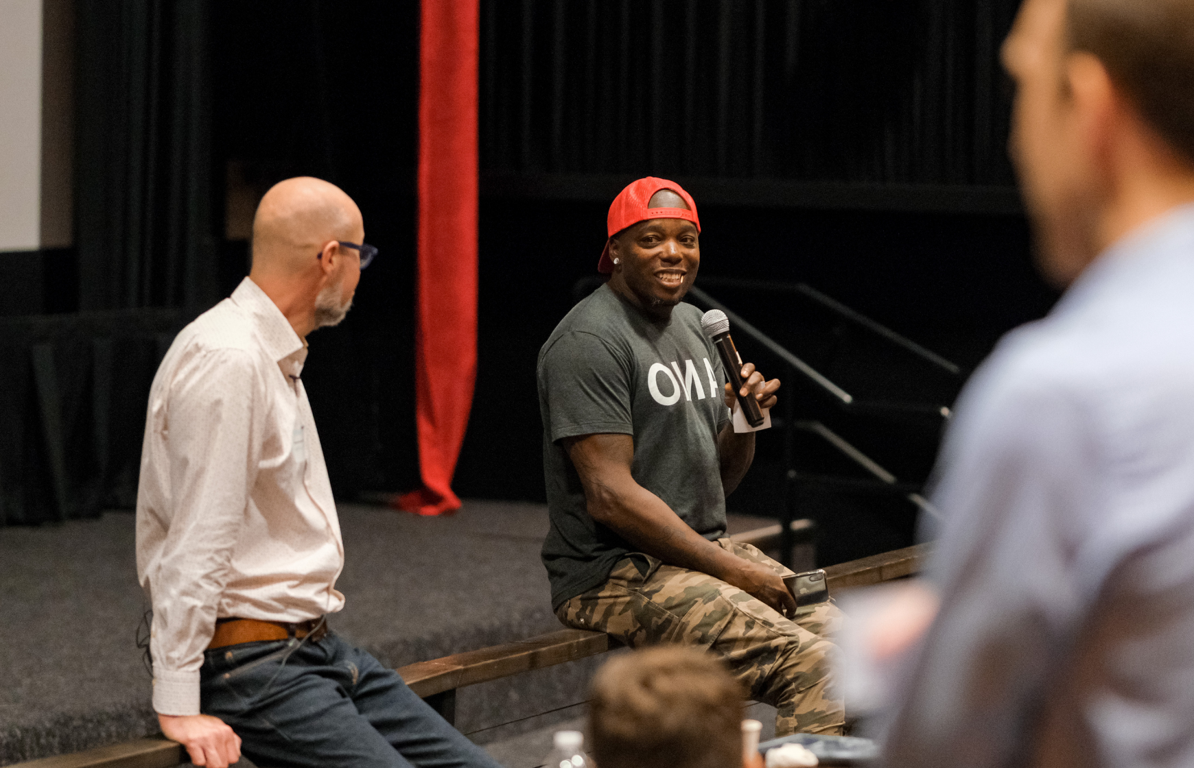 Man looks on as other man speaks into a microphone